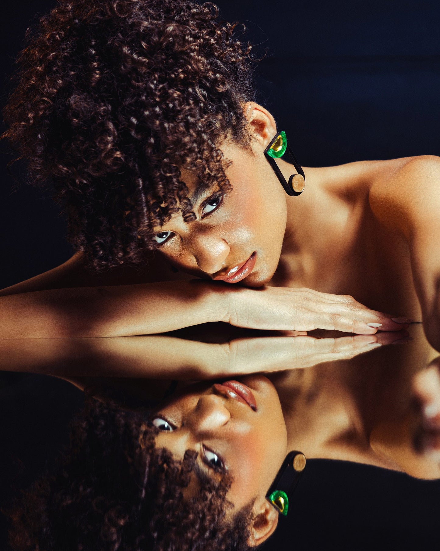 Woman with curly hair and green earrings looking at her reflection in a mirror.